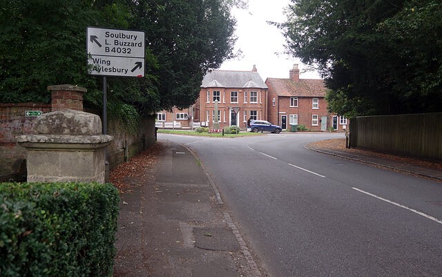 英国乡村路口实拍：白底黑字路牌标注 Soulbury / Leighton Buzzard / B4032 与 Wing / Aylesbury，左侧石砌门柱带球饰与弧形矮墙，远处为红砖坡顶宅邸，一辆深色旅行车停在路缘
