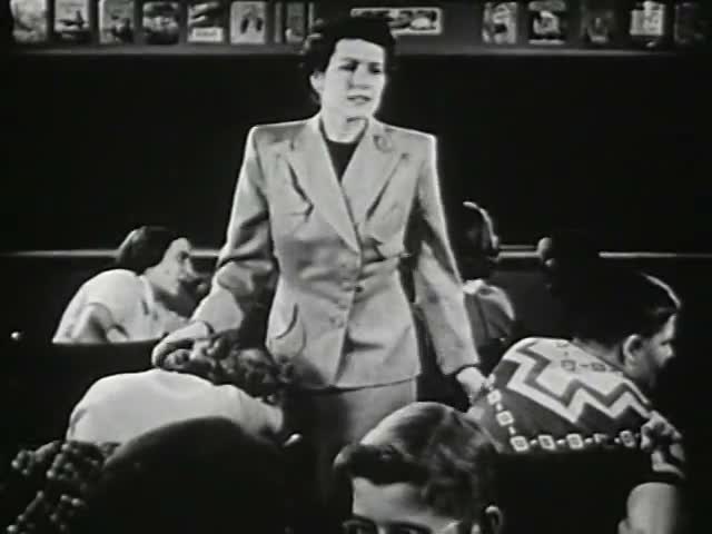 源片段第 0 秒采样帧：站立的女性（老师）面对坐着的孩子们讲话（黑白画面）