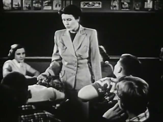 源片段第 4 秒采样帧：站立的女性（老师）面对坐着的孩子们讲话（黑白画面）